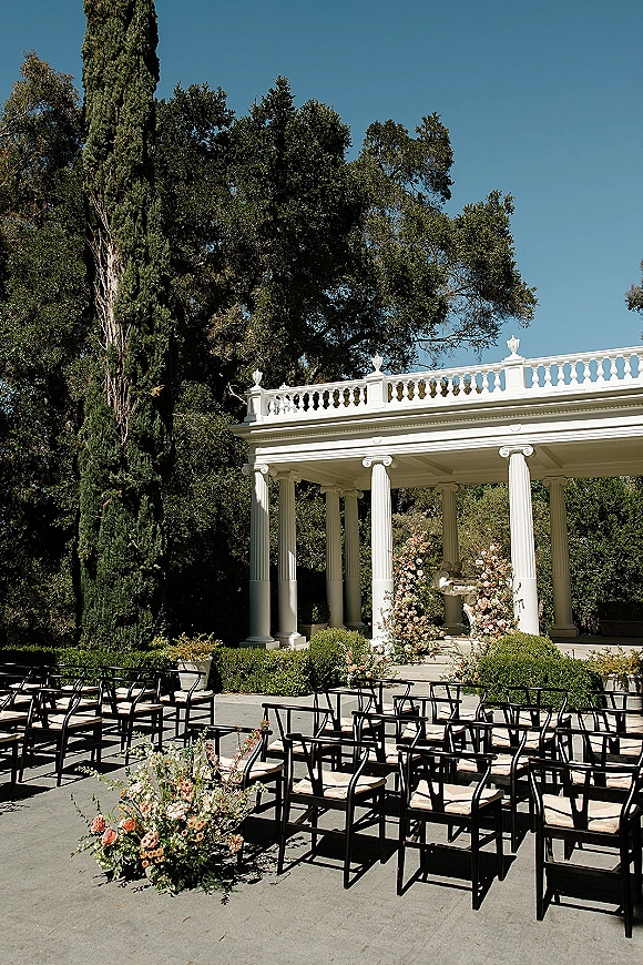 Ceremony setup with black ceremony chairs and blush floral aisle arrangements facing a white columned pavilion under tall trees and blue sky