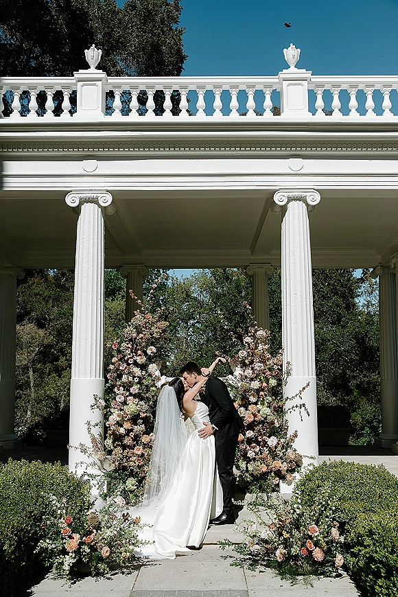 Wedding kiss as bride and groom kissing beneath a floral arch at a white column gazebo, her veil trailing along the stone walkway