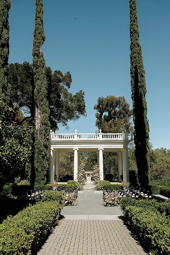 Ceremony setup with outdoor ceremony setup, stone aisle lined with blush and white florals leading to white columns under a sunny garden sky