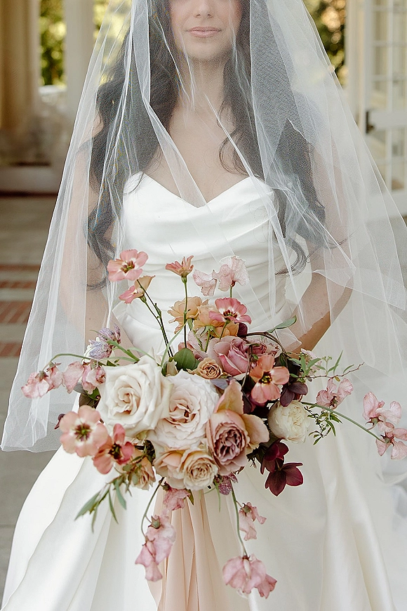 Bridal portrait of a bride with veil over her face, holding a blush rose bouquet with ribbon by an outdoor doorway with columns and steps