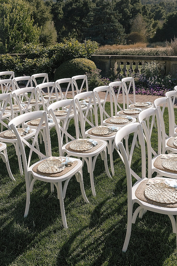 Ceremony seating with outdoor wedding chairs, white crossback chairs topped with straw hats and light blue ribbon bows on a garden lawn