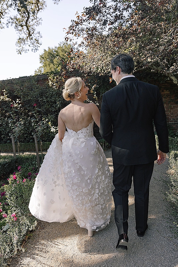 Couple portrait of bride and groom walking down a sunlit garden path, bride lifting her lace train beside him in a tuxedo