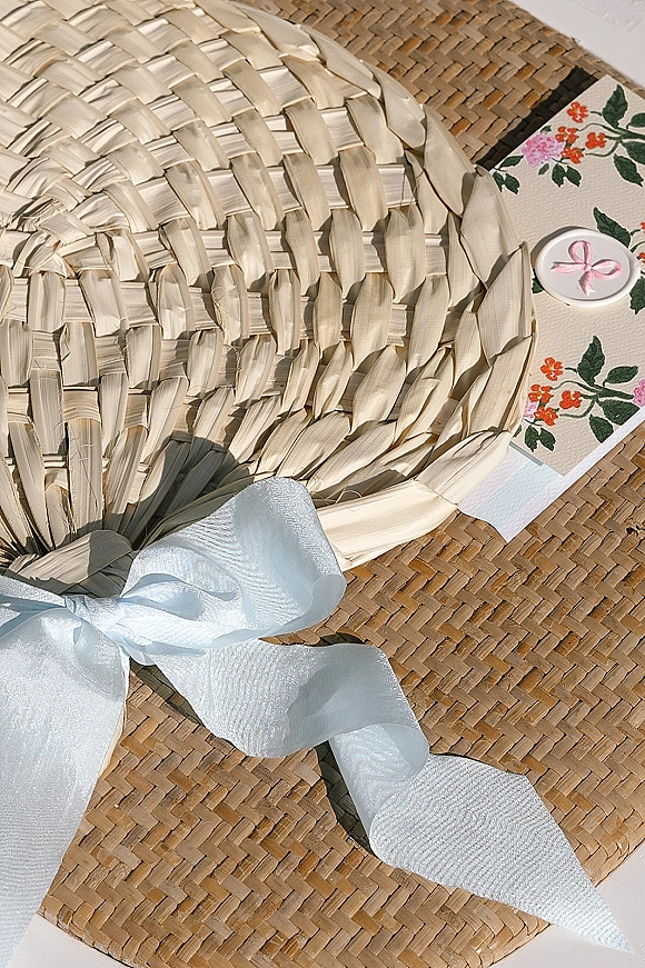Wedding invitation suite with floral wedding invitations, light blue ribbon bow and pink wax seal on a woven rattan surface with straw hat