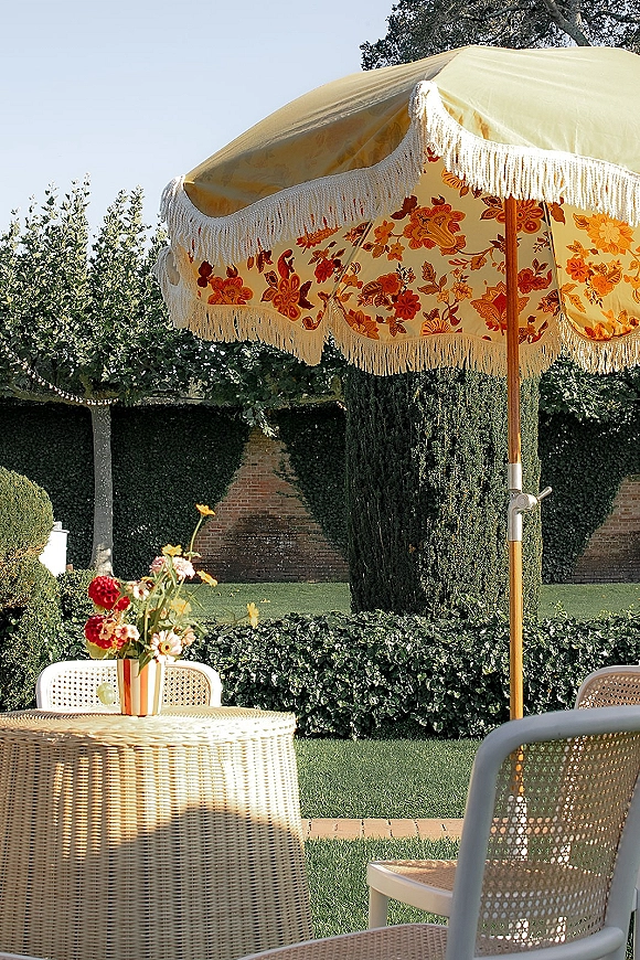 Outdoor lounge setup for wedding cocktail hour seating with a fringed floral patio umbrella, cane chairs, and striped vase on a garden lawn