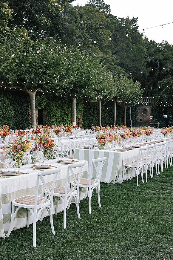 Outdoor reception tablescape with long banquet table wedding details, striped linens, floral centerpieces, white cross back chairs under string lights