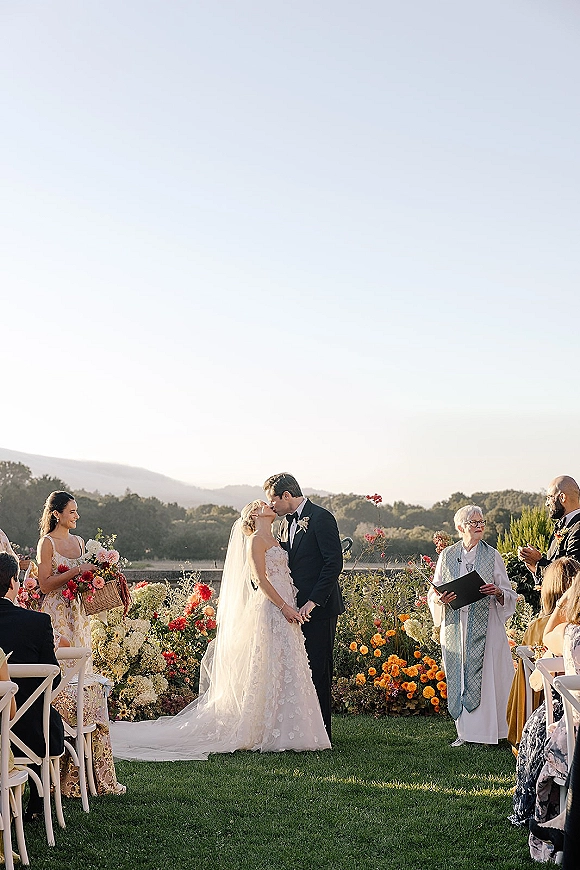 Ceremony kiss at an outdoor wedding ceremony under a flowered arch, bride in lace gown and veil with mountains behind guests