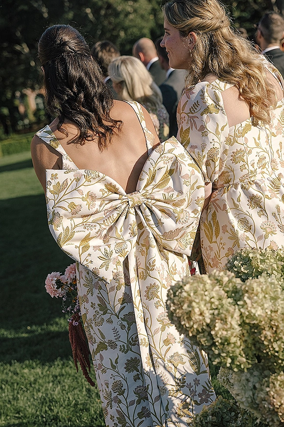 Bridesmaid dresses with an oversized bow back and puff sleeves, floral fabric and hydrangea bouquets on a garden lawn among guests