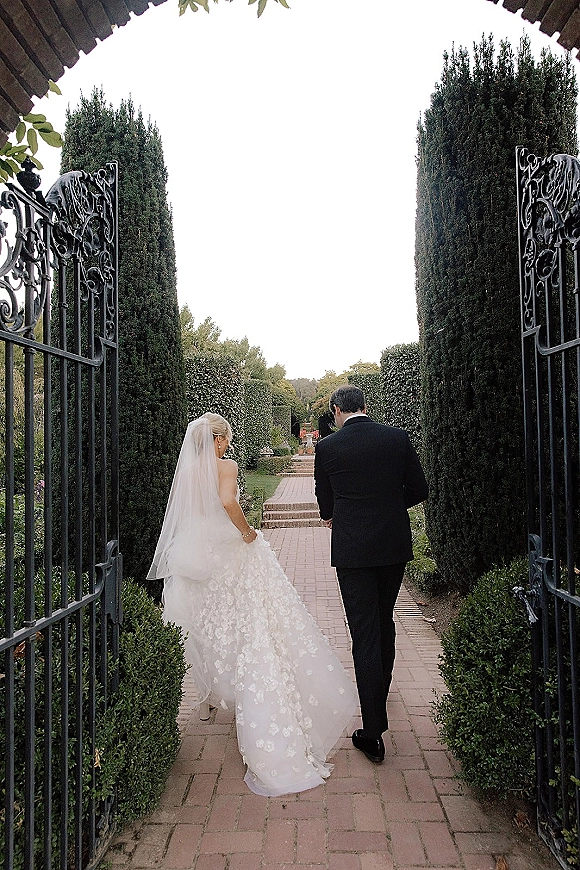 Wedding couple walking through wrought iron gates, bride lifting her dress train and veil beside groom in black tuxedo on a brick garden path