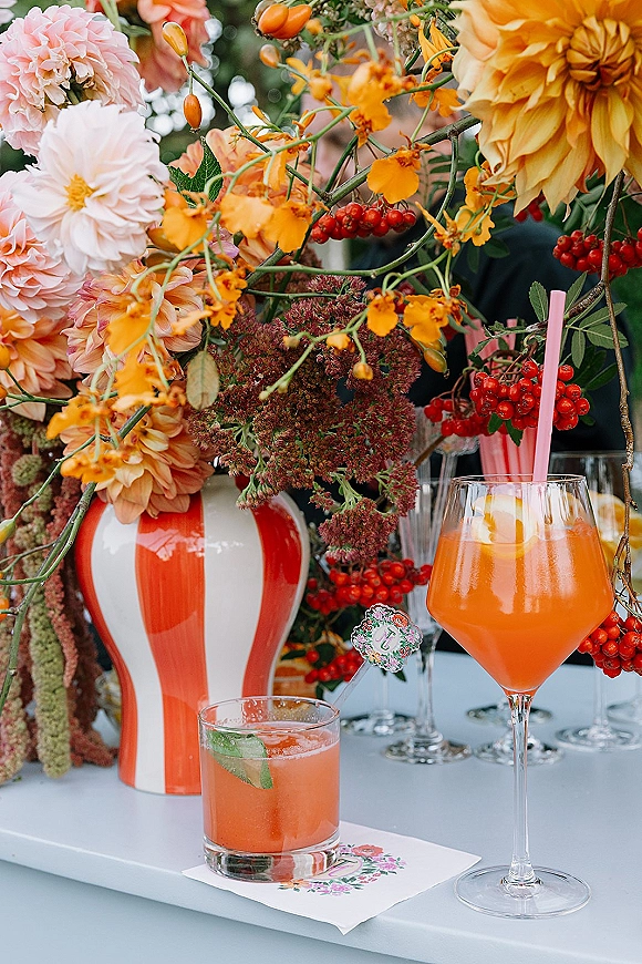 Wedding cocktail bar with signature wedding cocktails, pink straws and citrus garnishes beside a dahlia arrangement on a counter with blurred guests