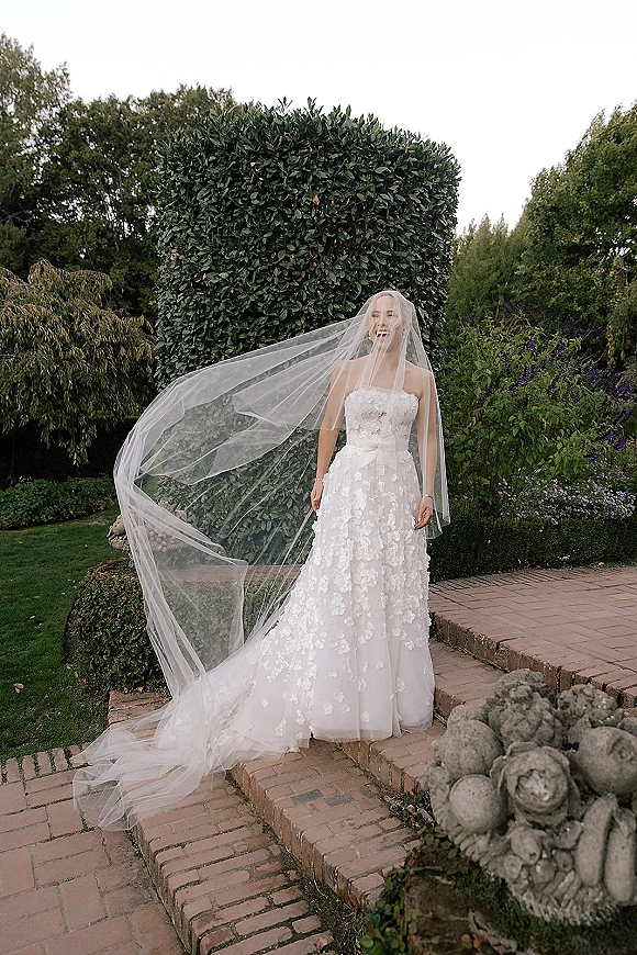 Bridal portrait of a bride with long veil blowing in the wind, wearing a strapless floral appliqué gown on brick garden steps by hedges
