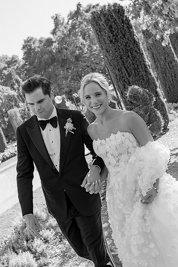 Couple portrait in a black and white wedding portrait, bride and groom holding hands on a garden pathway with tall cypress trees behind