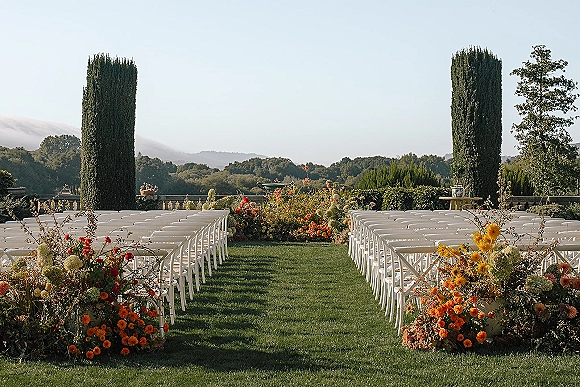 Ceremony setup for an outdoor wedding ceremony with white folding chairs and floral-lined aisle on a lawn terrace with cypress trees and hills
