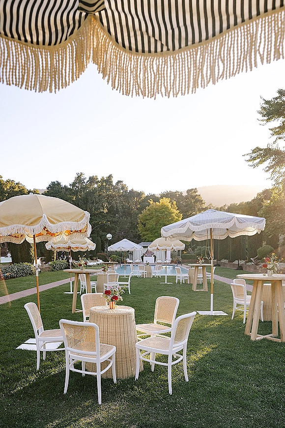 Cocktail hour setup with outdoor cocktail hour seating, striped patio umbrellas, cane chairs, and floral-topped tables by the pool lawn
