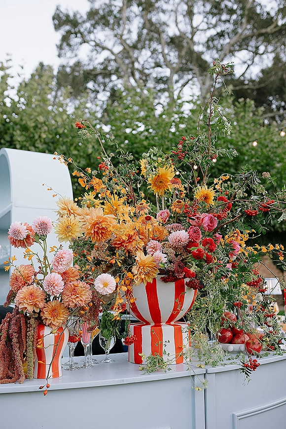 Reception tablescape with colorful wedding tablescape florals in striped pedestal vases, apples, wine glasses and string lights under trees