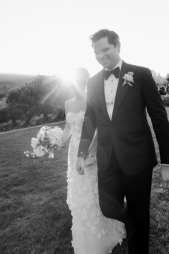 Wedding couple portrait of bride and groom walking hand in hand, her strapless lace gown and bouquet, with sun flare over lawn and hills