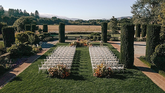 Outdoor ceremony setup with white crossback chairs and aisle runner, framed by garden hedges, cypress trees, and distant hills