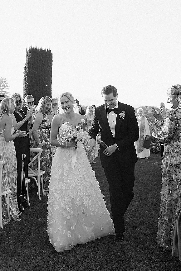 Wedding recessional as bride and groom walk down the aisle, bouquet and veil flowing, guests cheering on an outdoor lawn under trees