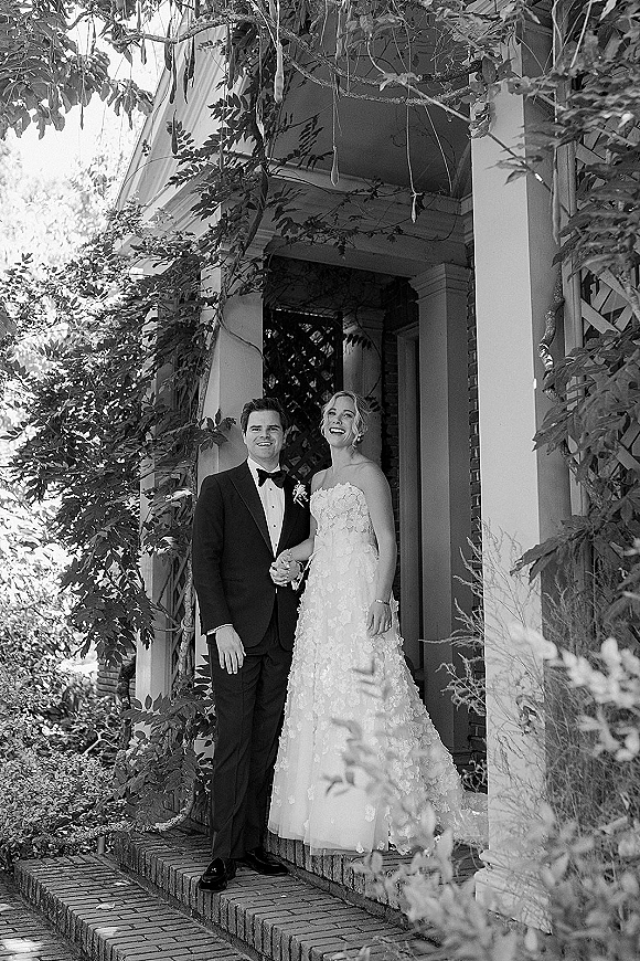Couple portrait in a black and white wedding portrait, bride in strapless lace gown and groom in tux holding hands on ivy-covered porch steps
