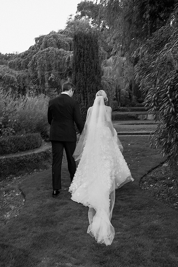 Wedding couple portrait of bride and groom walking away, her veil and dress train flowing up stone steps along a garden path