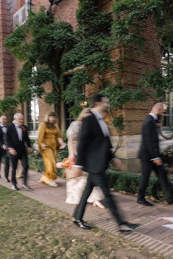 Wedding guests arriving in formal suits and bow ties, walking past a brick building with leafy trees and windows along the sidewalk