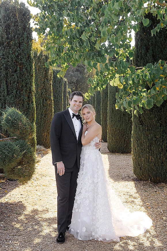 Couple portrait of bride in strapless floral appliqué gown and veil leaning on groom in black tuxedo along a sunlit garden path