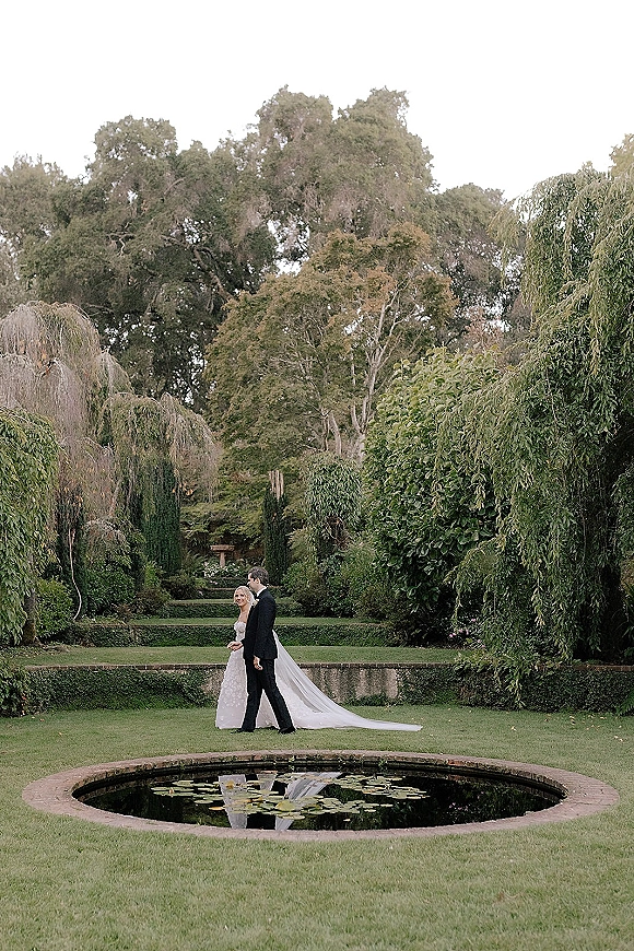 Couple portrait of bride and groom walking in outdoor wedding portraits, her long veil and dress train flowing by a reflecting pool with lily pads