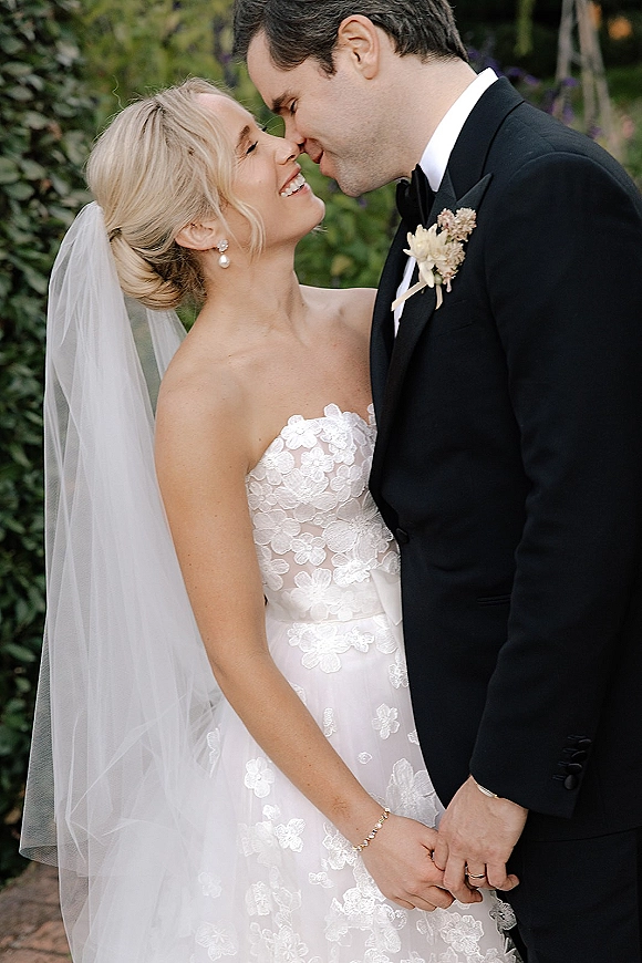 Wedding couple portrait of bride and groom close up touching foreheads, her veil and pearl earrings beside his black tuxedo amid garden greenery