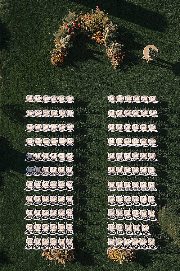 Outdoor ceremony setup with an aerial wedding ceremony layout showing white chairs in rows facing a curved floral arch on a sunlit lawn