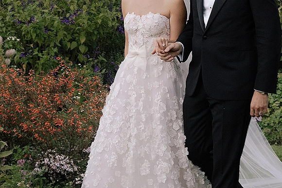 Couple portrait of bride and groom holding hands, showing her wedding ring and lace gown bow detail against garden greenery and flowers