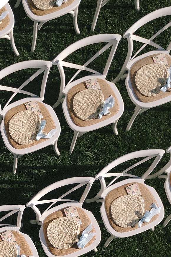 Ceremony chair decor with white wooden chairs, woven straw fans and blue ribbon bows, plus floral paper programs on a green lawn
