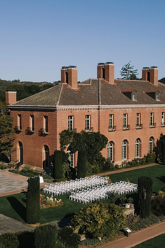 Ceremony setup with outdoor ceremony seating, rows of white chairs flanking a paved aisle on a lawn beside a brick building under blue sky