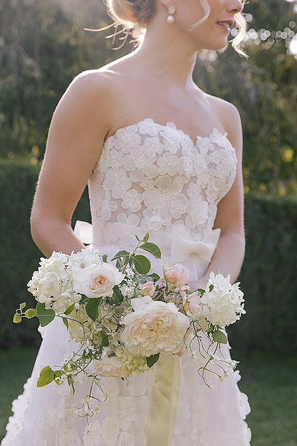 Bridal portrait of a bride holding bouquet of white and blush roses with greenery, in strapless lace dress in a sunlit garden bokeh background