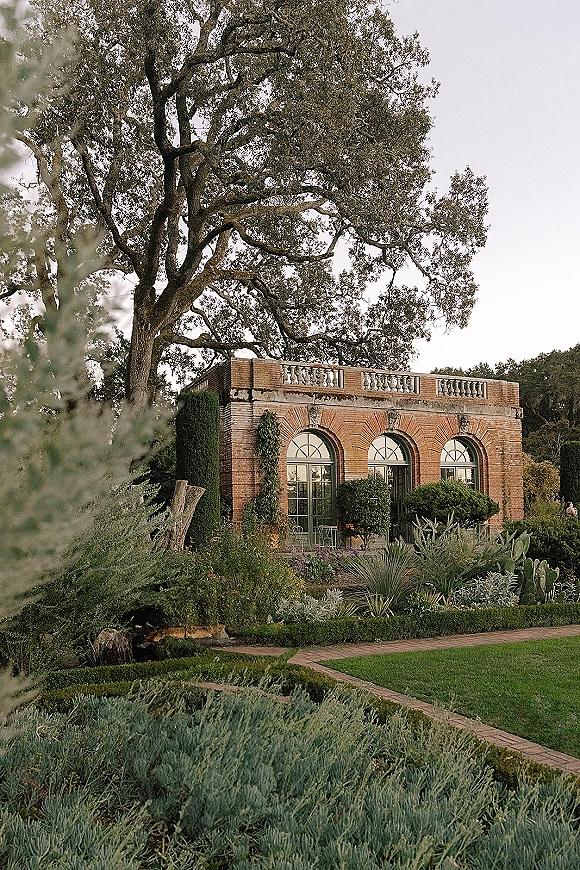 Garden wedding venue with brick building and arched windows, terrace balustrade, and manicured hedges lining a lawn path under trees