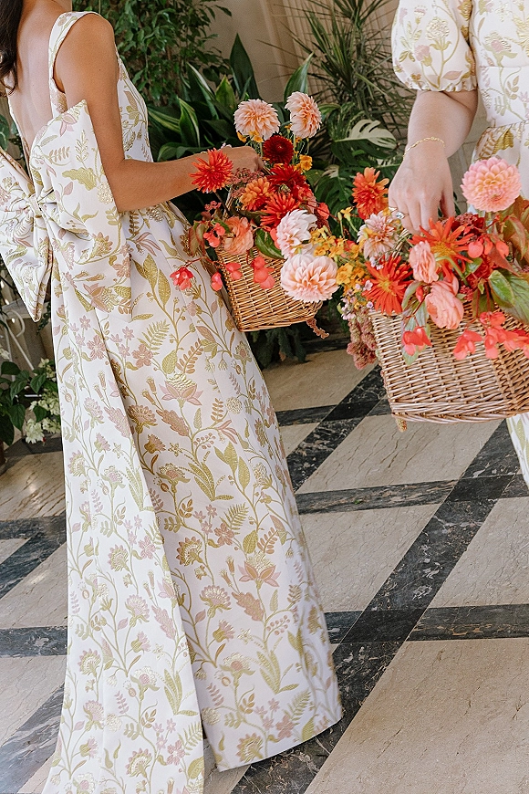 Bridesmaid bouquet in a wicker basket bouquet bursting with colorful dahlias, held by a floral print puff sleeve dress with gold bracelet indoors