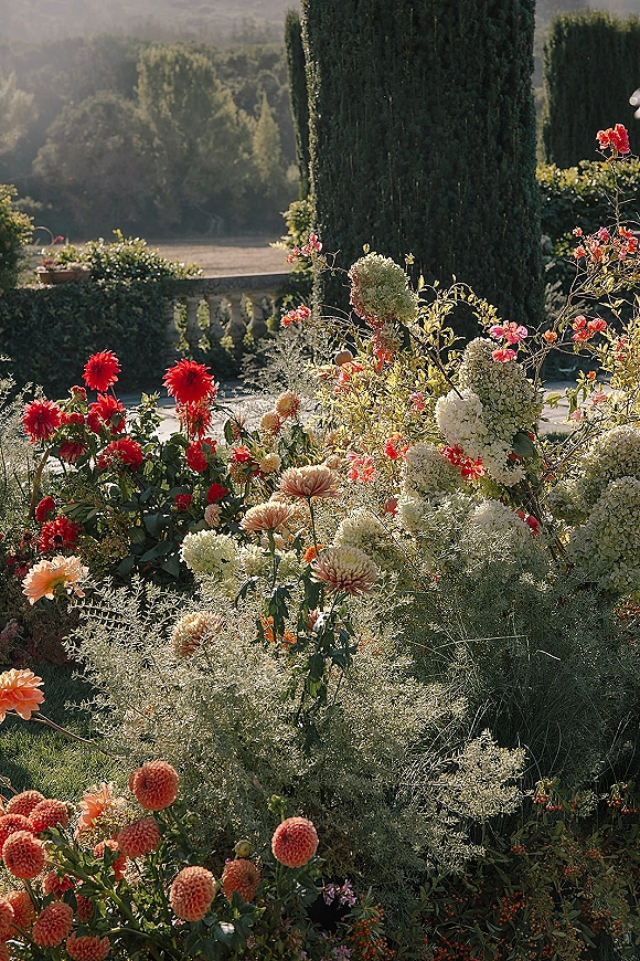 Wedding floral installation with hydrangeas, dahlias, roses, and greenery on a garden terrace beside a stone balustrade and cypress trees