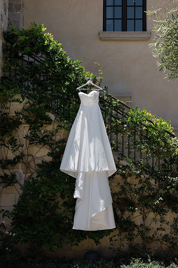 Wedding dress, strapless wedding dress hanging on a hanger over a black iron railing on an outdoor staircase with vines and stucco wall