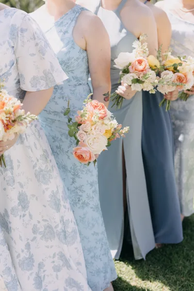 Bridesmaid dresses in blue bridesmaid dresses tones, mismatched with pastel bouquets, standing on a grassy lawn in daylight greenery
