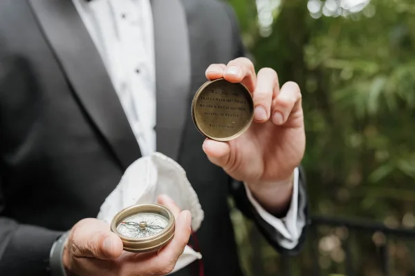 Engraved compass, a personalized groom gift, rests on white cloth as garden plants and an outdoor railing blur into green foliage in the background.