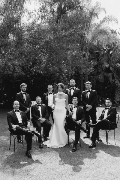 Wedding party portrait of a bride with groomsmen, some seated, holding martini glasses on a palm-lined garden lawn.