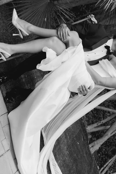 Groom carrying bride in a playful couple portrait, her flowing veil trailing as they pose on a palm-lined lawn beside sunlit stone pavers.