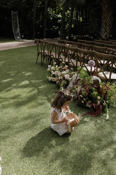 Flower girl moment as a flower girl with basket pauses on a grass lawn beneath garden trees and palm fronds in a serene garden setting.