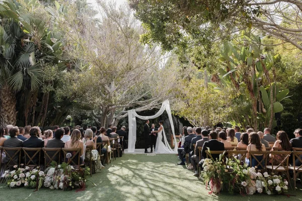 Ceremony moment at an outdoor wedding ceremony as the couple exchange vows under a simple draped arch amid tropical garden trees.