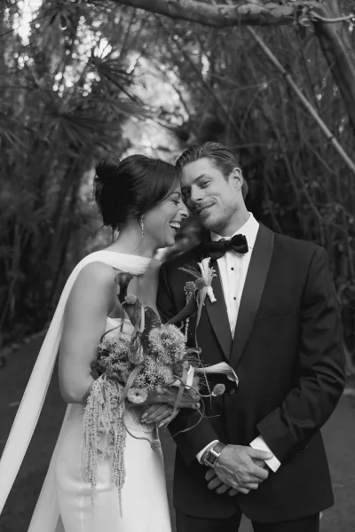 Couple portrait of bride and groom laughing, she cradles a sculptural bridal bouquet beneath arching garden branches along a leafy pathway.