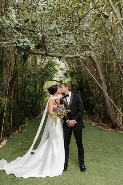 Wedding kiss portrait as a bride and groom kiss beneath a bamboo grove, her purple bouquet between them, natural light through leaves.