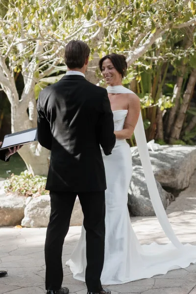 Ceremony moment during wedding vows: bride in a strapless satin column dress with a bridal cape, under garden trees and beside stone boulders.