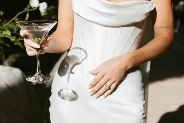 Wedding dress detail showcasing a satin bridal gown bodice as the bride holds a martini glass, set against sunlit garden greenery and soft shadows.