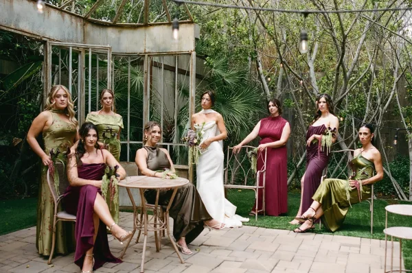 Bridal party portrait of a bride with bridesmaids holding bouquets in a garden beneath string lights by a greenhouse frame and palms on stone pavers