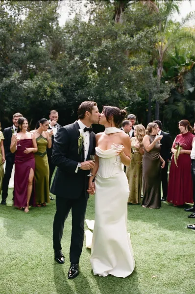 Wedding kiss portrait as the bride and groom kiss beneath garden palms, a candid moment with his boutonniere prominently in view.