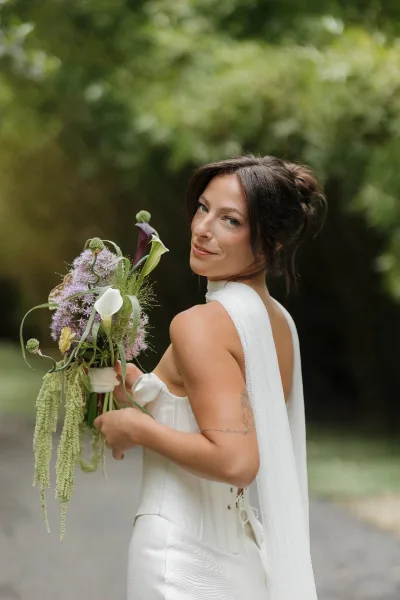 Bridal portrait of a bride holding bouquet in an over-the-shoulder pose, set among garden trees with a pathway behind.