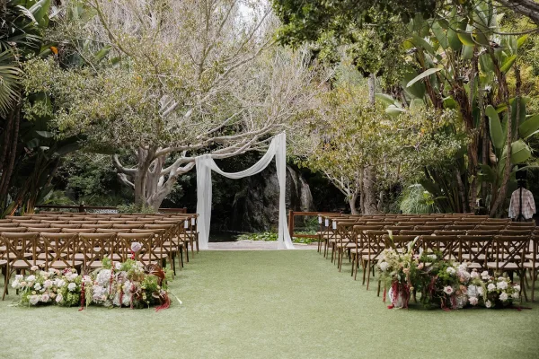 Ceremony setup with elegant garden ceremony decor featuring draped fabric, by a pond on a lawn bordered by tropical plants and garden trees.